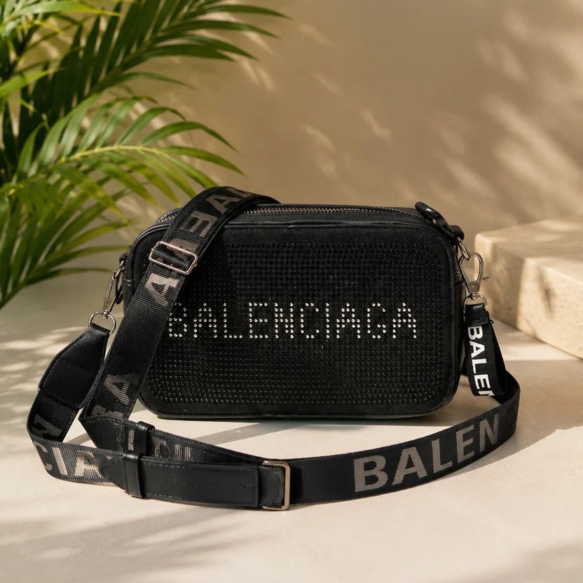 A black Balenciaga crossbody bag with a rhinestone-embellished front and a wide black strap featuring white BALENCIAGA lettering, styled on a beige surface with palm leaves and soft shadows.