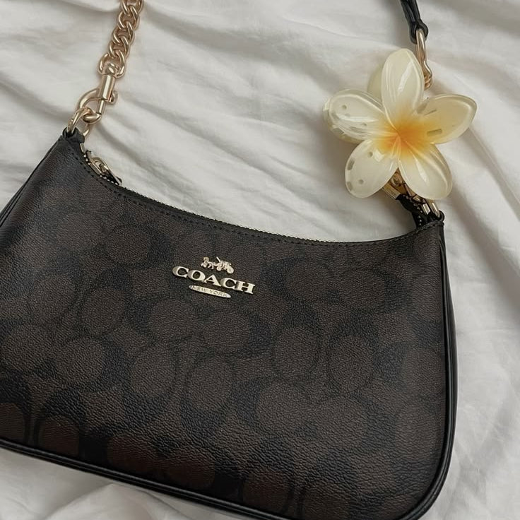 A Coach shoulder bag in the signature dark brown monogram canvas, featuring a black leather handle with a gold-tone chain link, a gold-tone logo plate, and a decorative white plumeria flower charm.